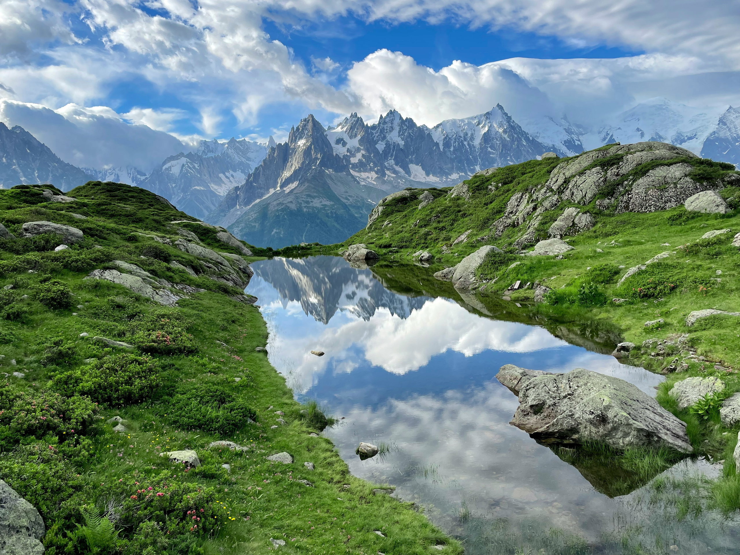 The reflection of the Aiguilles de Chamonix and the Mont Blanc massif in the calm waters of the Lacs des Chéserys, framed by green alpine meadows.