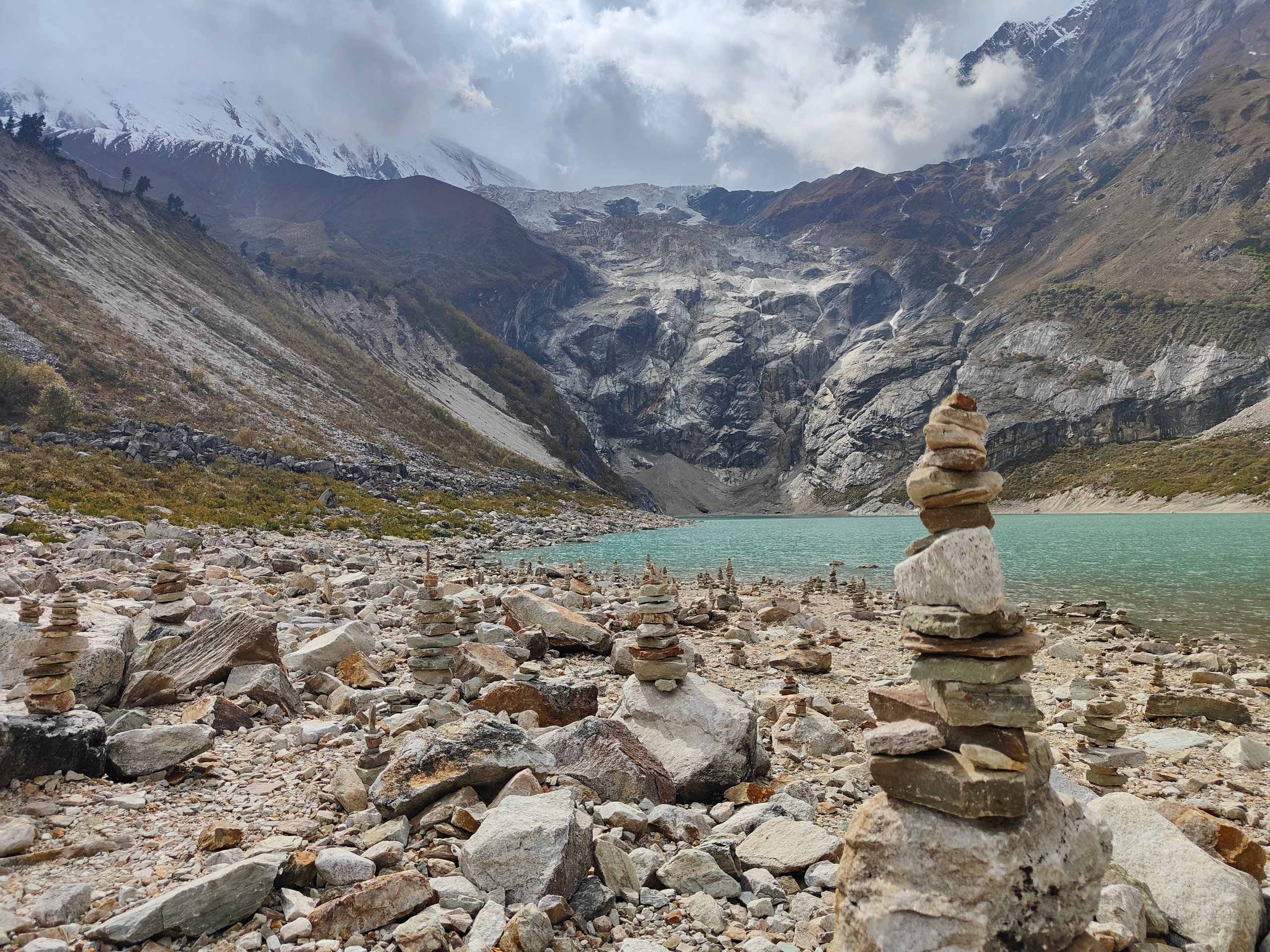 Stone cairns on a rocky shore at Birendra Tal glacial lake, with the snowy Manaslu peaks in the background