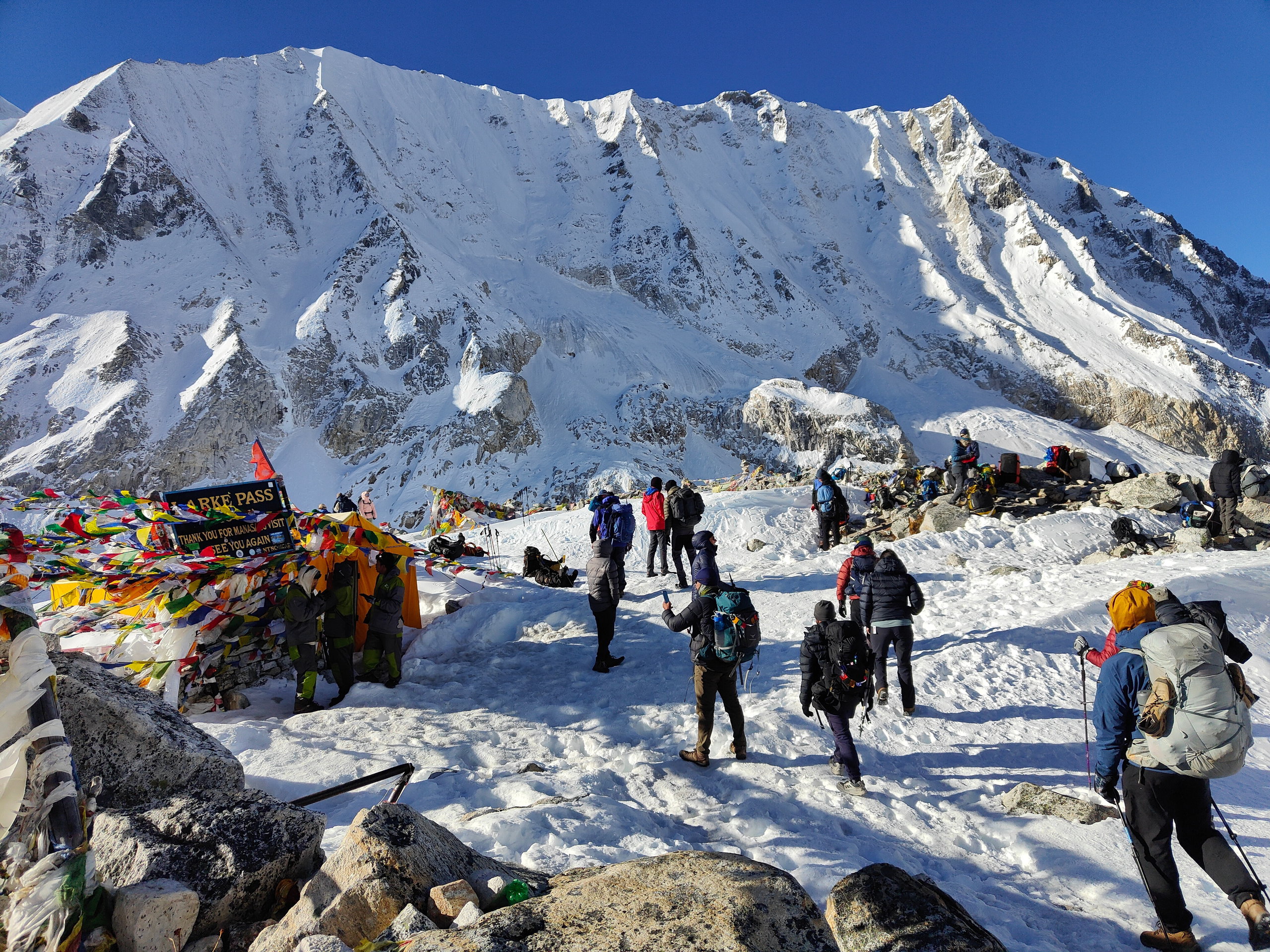Larkya La pass with a sign at 5106 meters, colorful prayer flags, and trekkers on snowy ground facing the mountain
