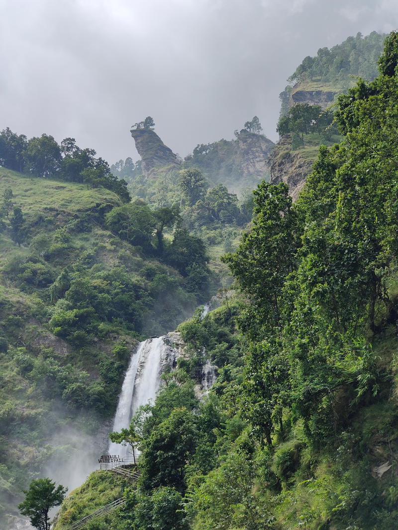 Waterfall flowing down a rocky cliff in tropical forest, with an observation platform on the left and misty mountain ridges in the background