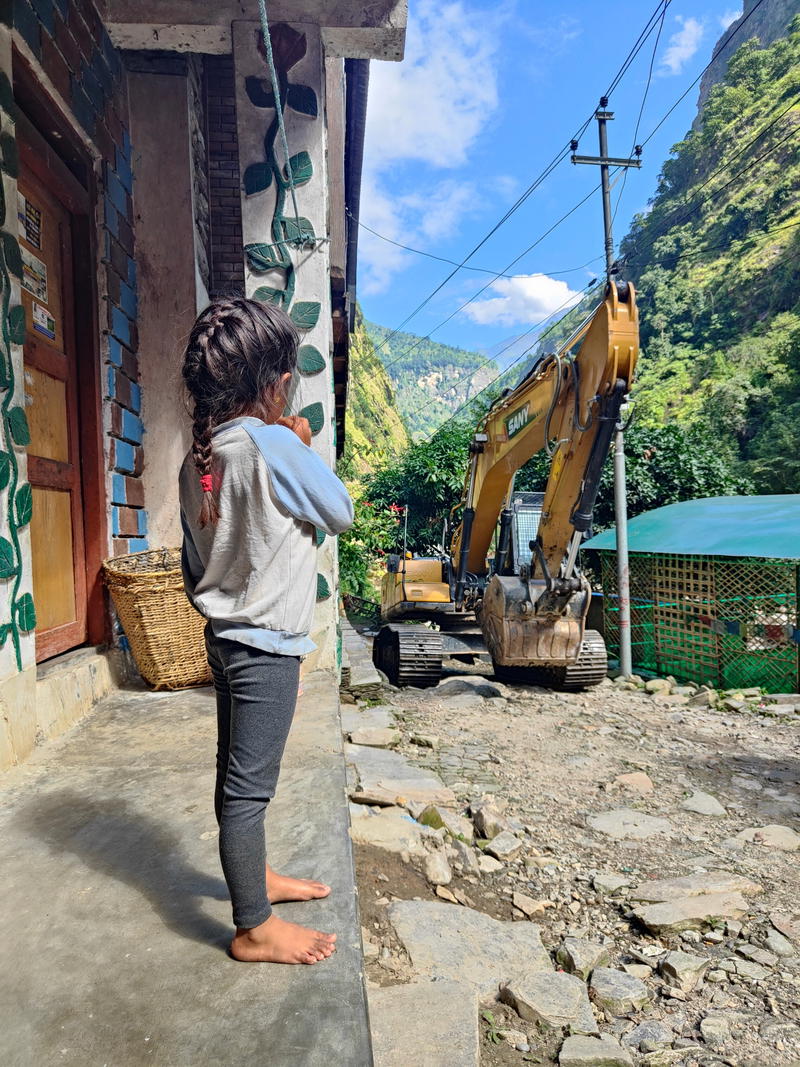 Child watching a yellow Sany excavator driving along a narrow path between houses in a mountain village, with wooded slopes in the background