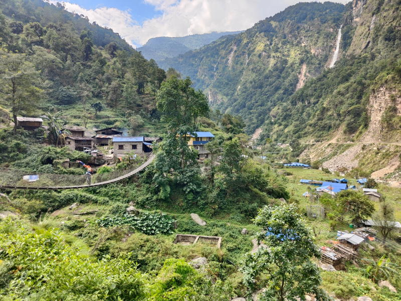 Narrow suspension bridge crossing a green valley toward a village of stone houses with blue roofs, with wooded mountains and a waterfall in the background
