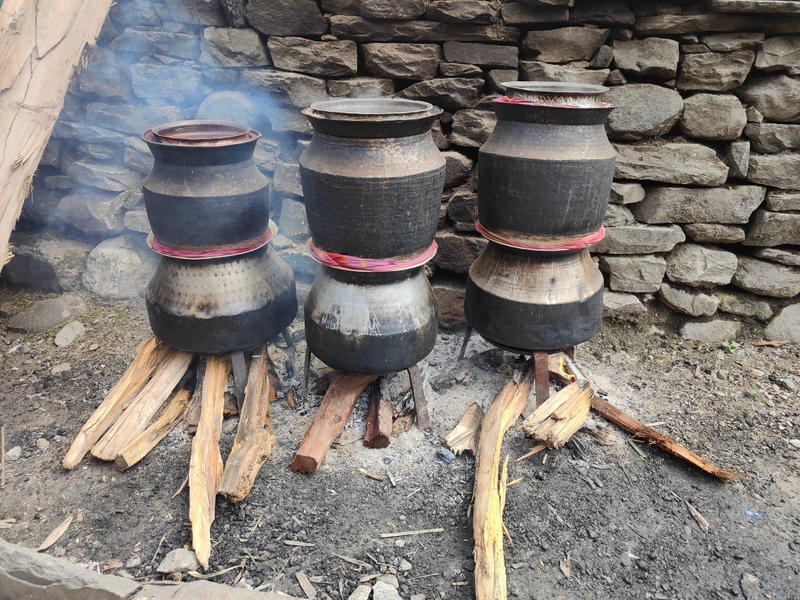 Three traditional stills made from blackened metal pots heating over wood fires against a dry-stone wall to produce Raksi, the local alcohol.