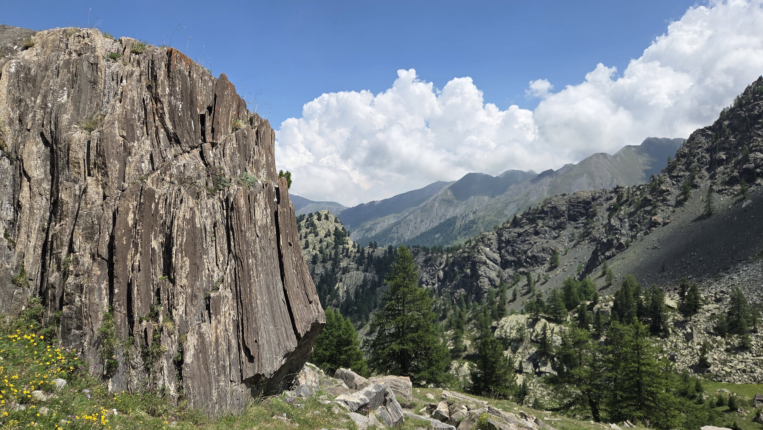 A large, vertically striated rock formation on a mountain trail with pine trees and alpine peaks in the background under a blue sky