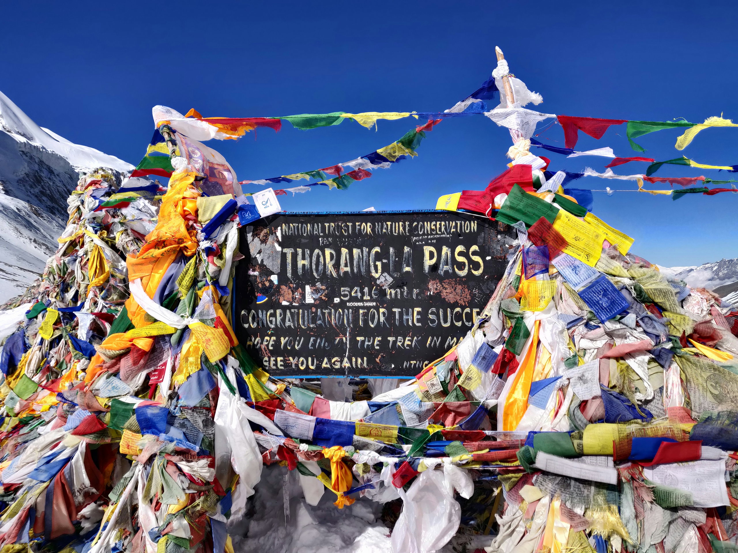 Sign at the Thorong La Pass at 5,416 m, surrounded by prayer flags and snow-capped mountains.