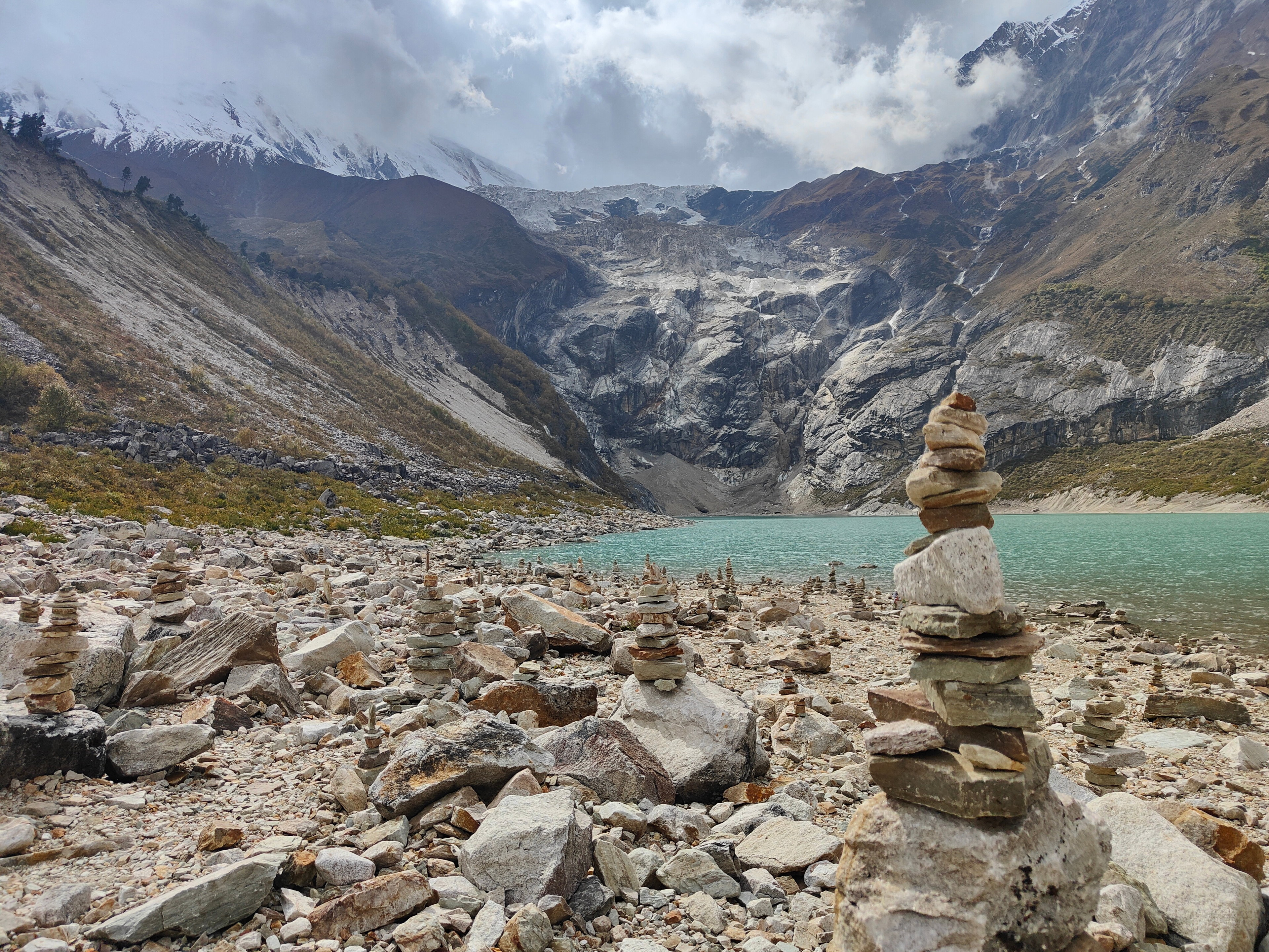Stone cairns on a rocky shore at Birendra Tal glacial lake, with the snowy Manaslu peaks in the background