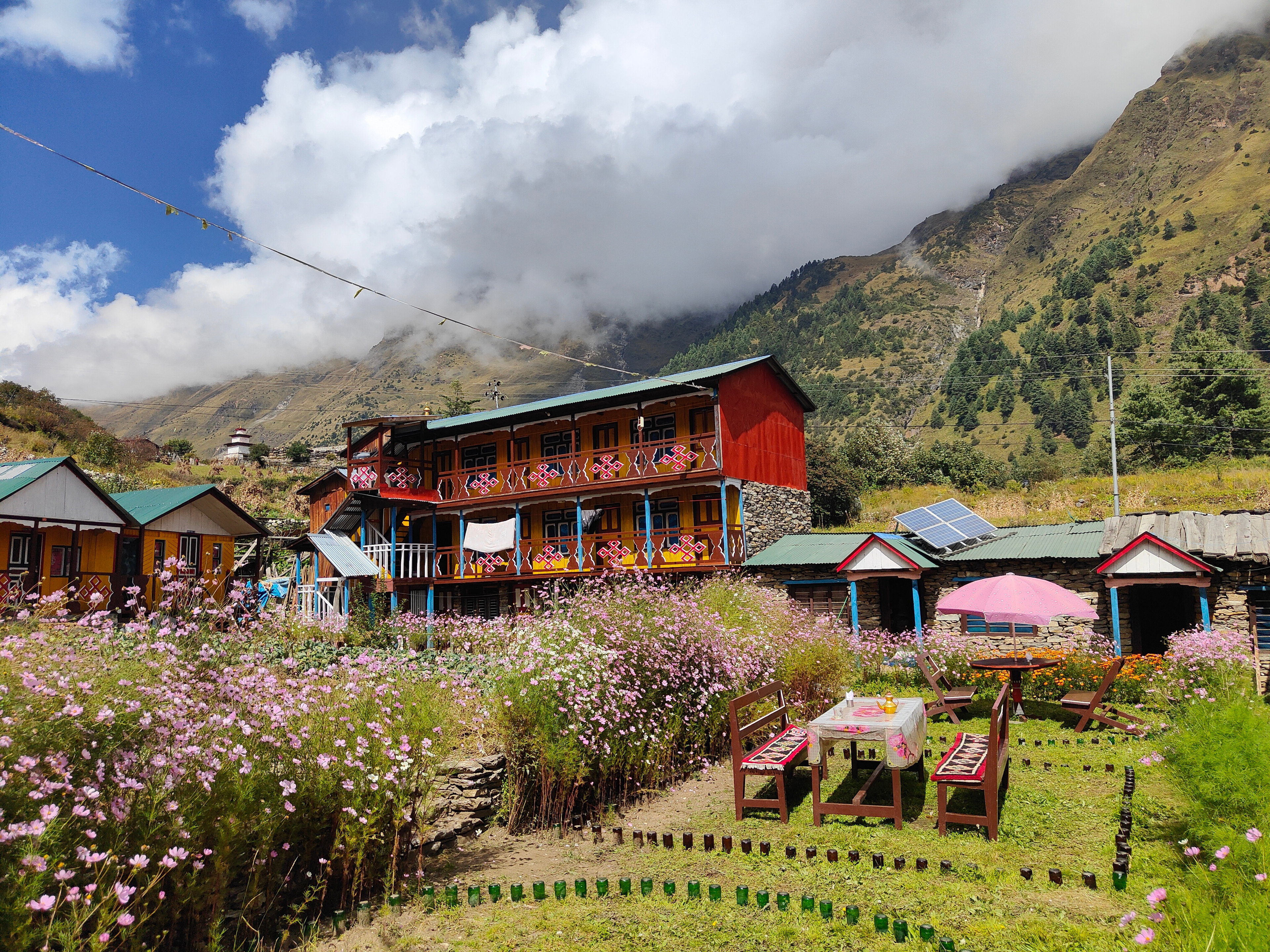 Two-story yellow and red lodge behind a garden of pink flowers, with a terrace and solar panels under the clouds