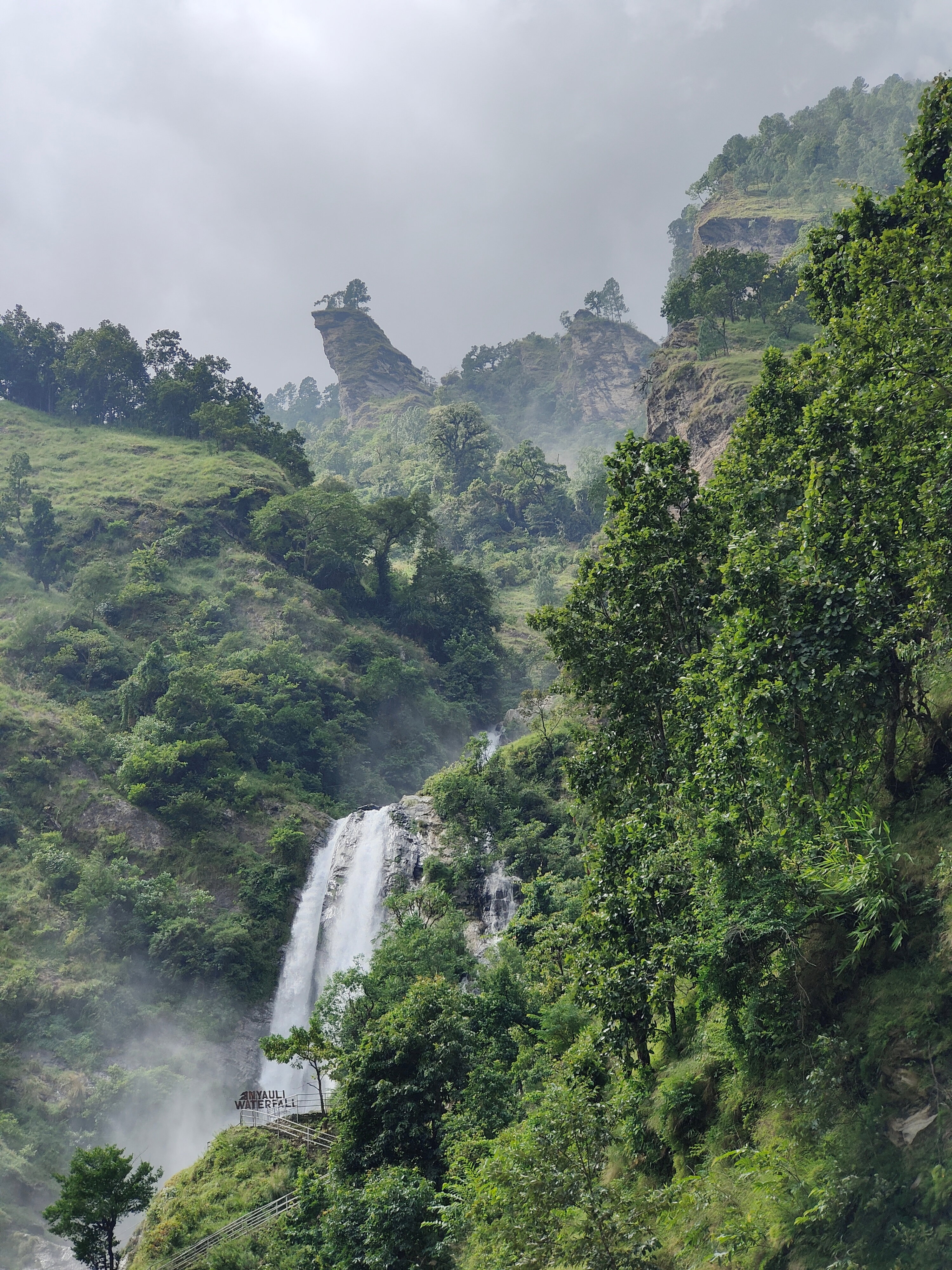 Waterfall flowing down a rocky cliff in tropical forest, with an observation platform on the left and misty mountain ridges in the background